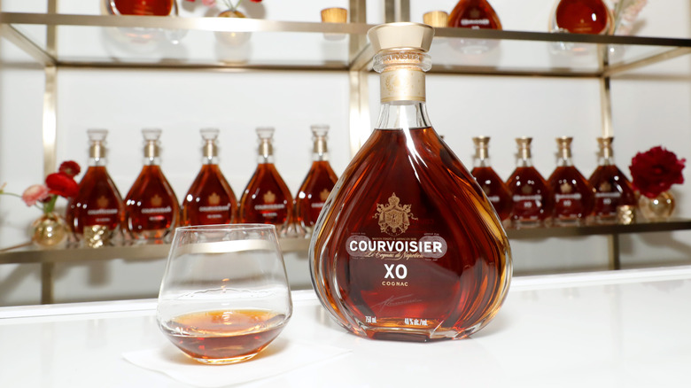 A bottle of Courvoisier XO on a bar next to a glass with bottles of the XO in a stored behind it