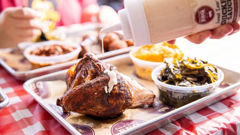 Drizzling sauce from a bottle onto BBQ chicken on tray with sides