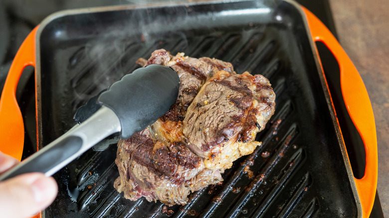 Person turning ribeye steak over on a cast iron grill pan