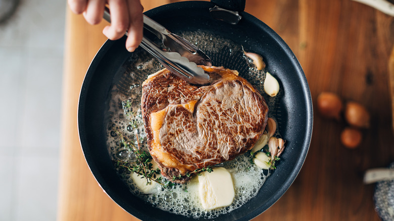 A hand with tongs holding a steak cooking in a pan with butter and herbs