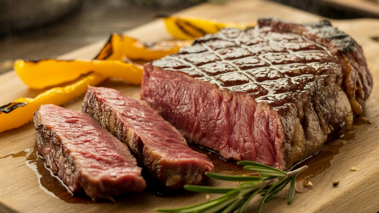A seared ribeye steak on a wooden board with grilled peppers alongside it