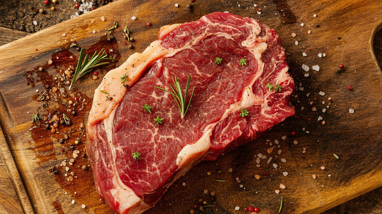 A raw ribeye steak on a wooden board with crushed salt and pepper flakes around it