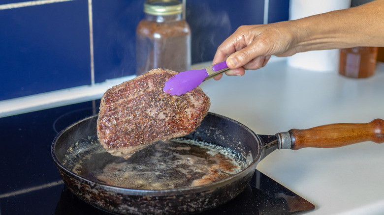 A hand holding a steak with tongs above a pan