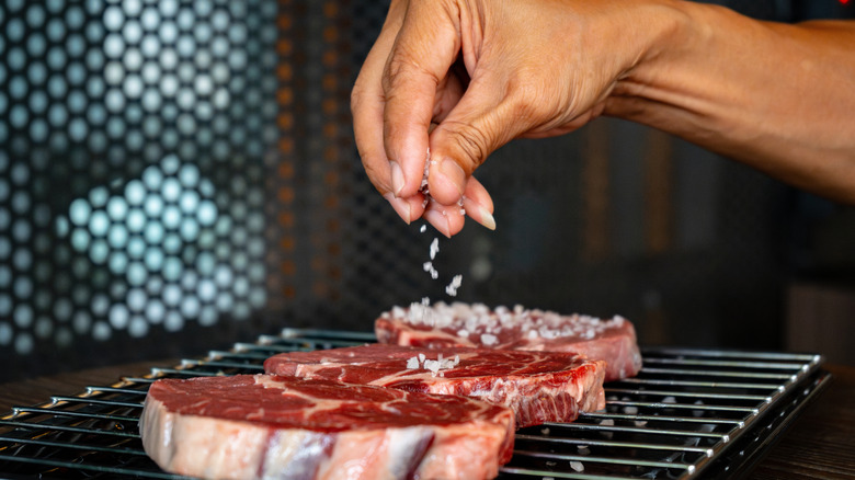 A hand adding salt to ribeyes on a grill