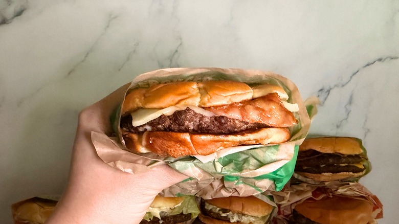 Hand holding Bacon Swiss Buttery Smashed Jack burger from Jack in the Box with burgers on marble surface in background