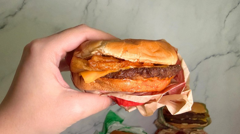Hand holding Hot Ones Sriracha Curly Fry burger from Jack in the Box, burgers on marble surface in background