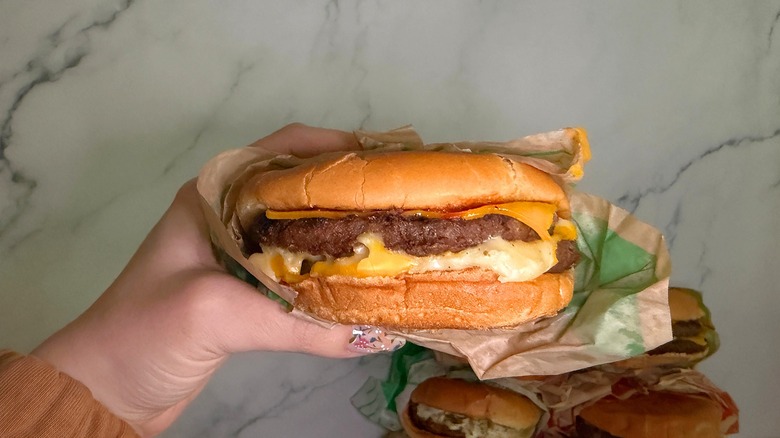 Hand holding Ultimate Cheeseburger from Jack in the Box with burgers on marble surface in background