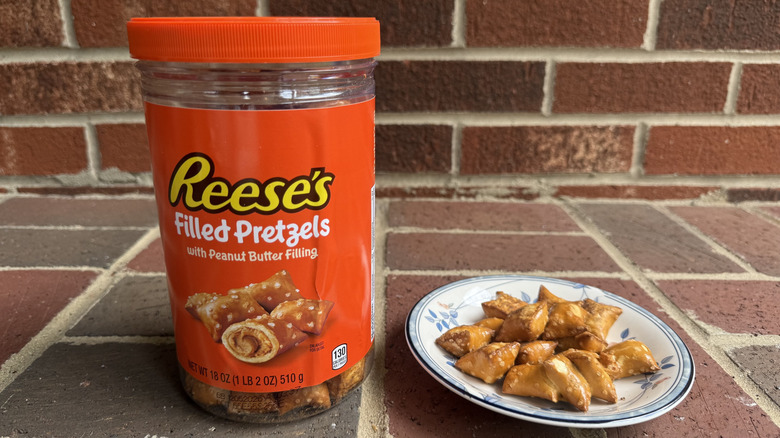 Medium plastic container of Reese's peanut butter-filled pretzels on brick surface, with some displayed on adjacent plate