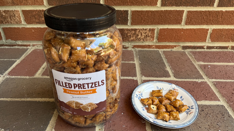 Tall plastic container of Amazon Grocery peanut butter-filled pretzels on brick surface, with some displayed on adjacent plate
