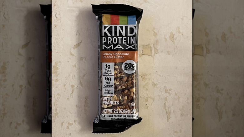 A Kind Protein Max crispy chocolate peanut butter flavor bar on a textured off-white surface