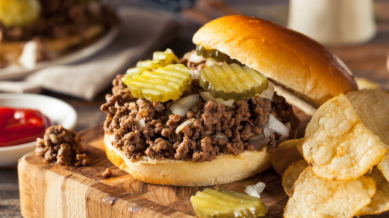 A Maid-Rite, Tavern, or loose meat sandwich on a chopping board next to some potato chips
