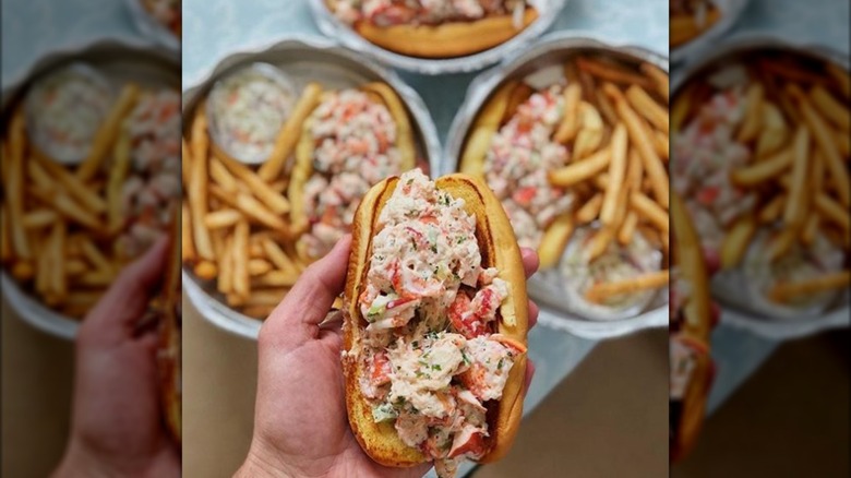 Hand holding a lobster roll above blurred images of more lobster rolls and fries dishes