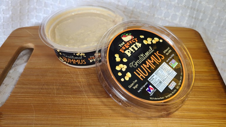 Open tub of The Perfect Pita hummus on wooden cutting board