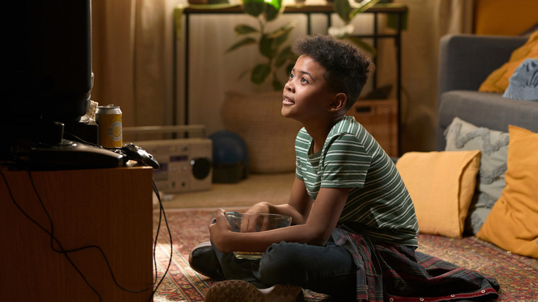 Kid sitting on floor in vintage-looking living room watching TV and eating popcorn