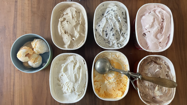 A bowl of ice cream and six open quarts of ice cream with a scoop