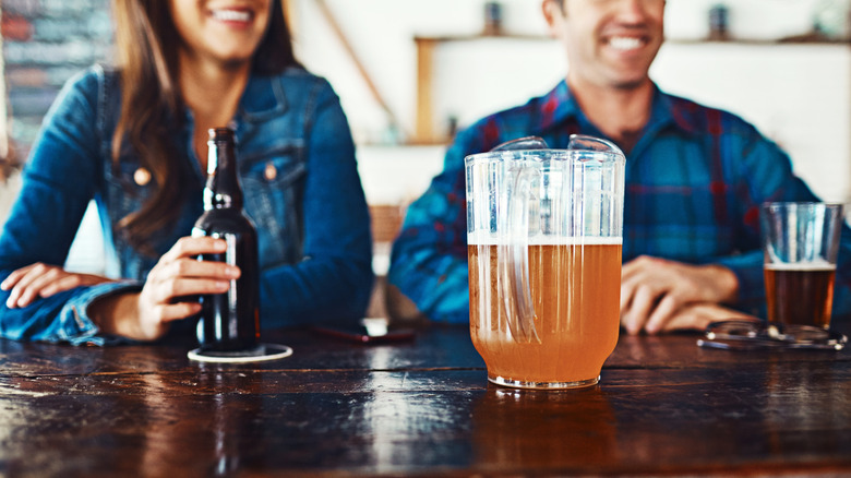Two people blurred in backrgound at bar with bottle, glass, and pitcher of beer