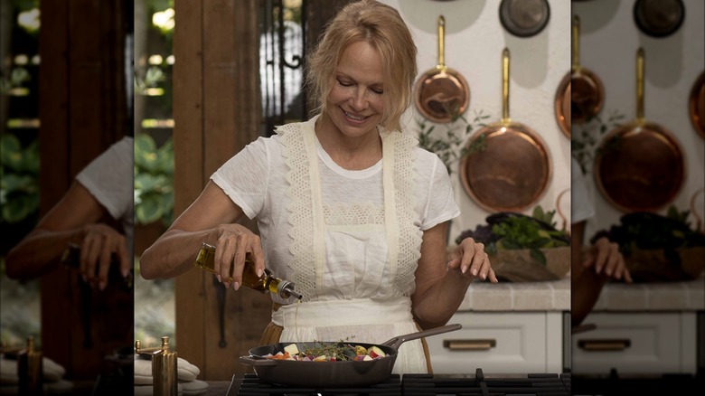 Pamela Anderson drizzling olive oil into a pan of vegetables and herbs