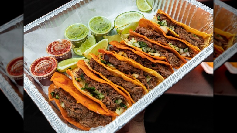 Eight birria tacos on tray with lime slices and salsa cups