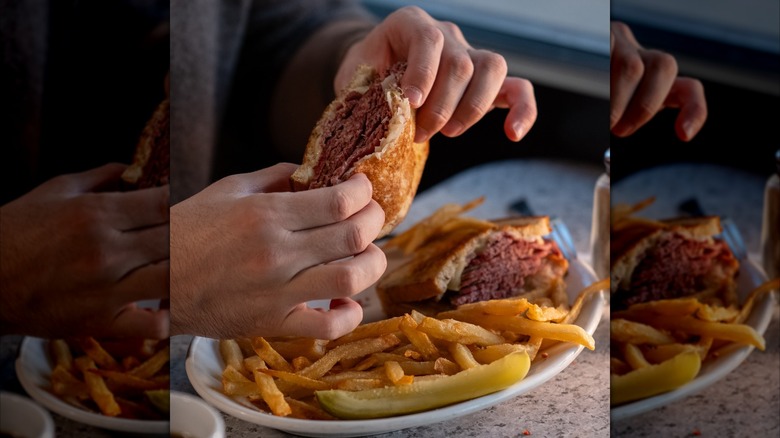 Hands holding a pastrami sandwich with plate of fries and pickle