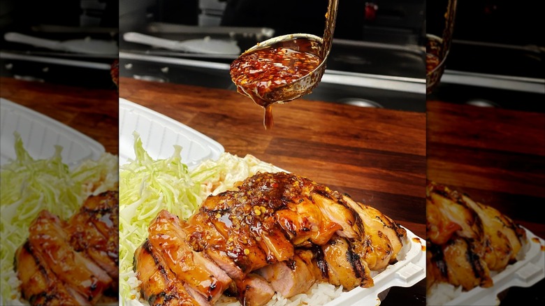 Spicy sauce being ladeled onto teriyaki chicken in a takeout container