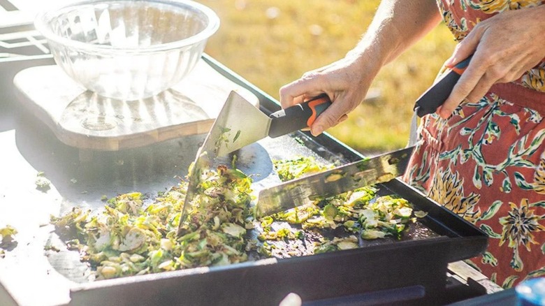 Person in a floral apron is using the Blackstone Pro Series Griddle Spatula Set to sauté Brussels sprouts