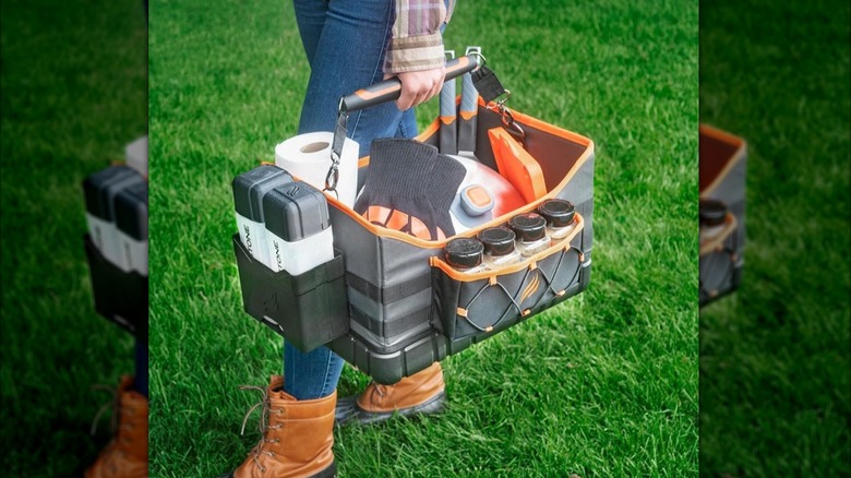 Blackstone Griddle Caddy full of squeeze bottles, tongs, gloves, spices, basting cover, and roll of paper towels being carried on a lawn