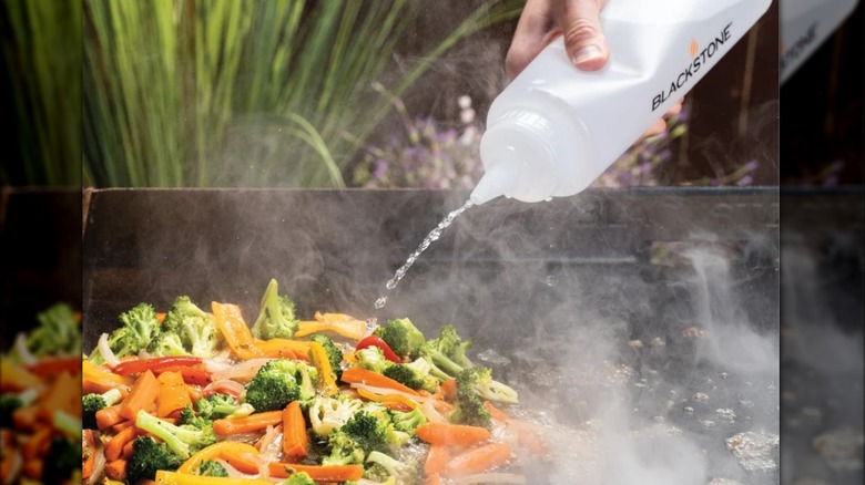 A hand squeezes water from a Blackstone Squeeze Bottle onto a variety of vegetables on a Blackstone Griddle