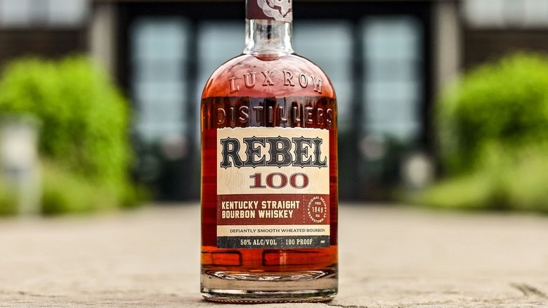 A bottle of Rebel 100 placed on the floor in front of a blurred Lux Row Distillery entrance