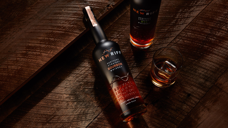 A bottle of New Riff Bottled-in-Bond laid on a wooden background next to another bottle and a filled glass