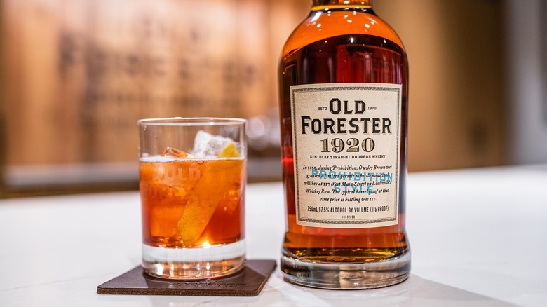 A bottle of Old Forester 1920 on a table next to a cocktail in a branded glass