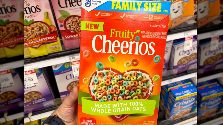 A hand holding a box of Fruity Cheerios in the store's cereal aisle.