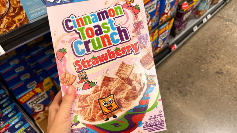 A hand holding a box of Cinnamon Toast Crunch Strawberry cereal.