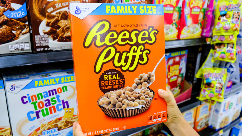A hand holding up a box of Reese's Puffs cereal in a store.