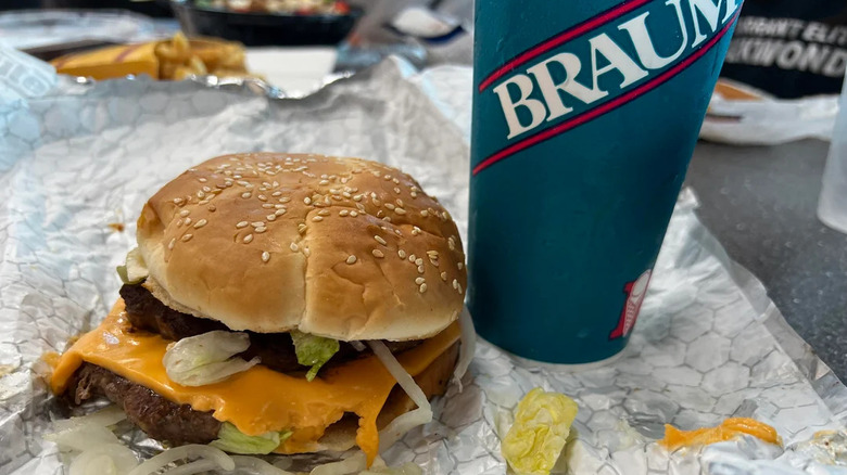 A Braum's cheeseburger with onions and lettuce scattered about it next to a drink