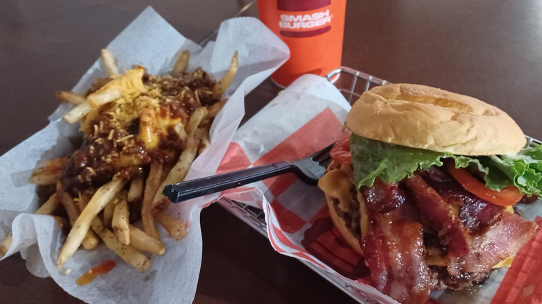 A Smashburger bacon cheeseburger next to chili cheese fries and a drink