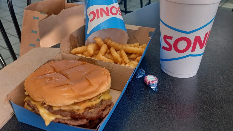 A cheeseburger and some crinkle cut fries in a box next to a drink from Sonic