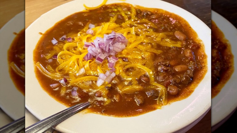 A bowl of Texas Roadhouse chili with a spoon in it, topped with melting cheese and red onion