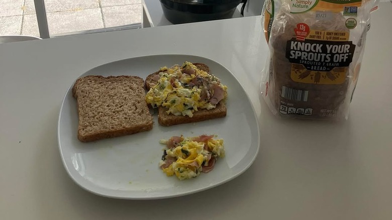 A breakfast sandwich with scrambled eggs and ham on sprouted grain bread sits on a plate beside a loaf of Simply Nature "Knock Your Sprouts Off" organic bread