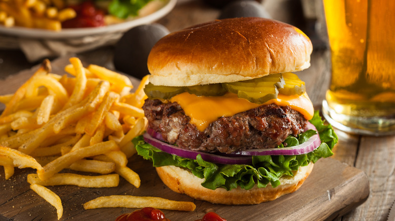 a bison cheeseburger on a wooden board with fries and a beer in the background