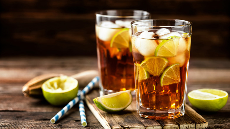 Two glasses of cola with ice and thin slices of lime next to halved limes and straws