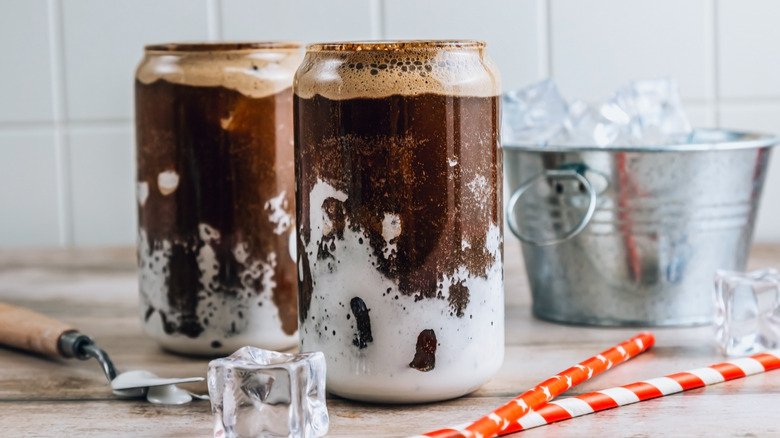 Two glasses of fluffy Coke drink with marshmallow fluff and cola next to striped straws and ice cubes