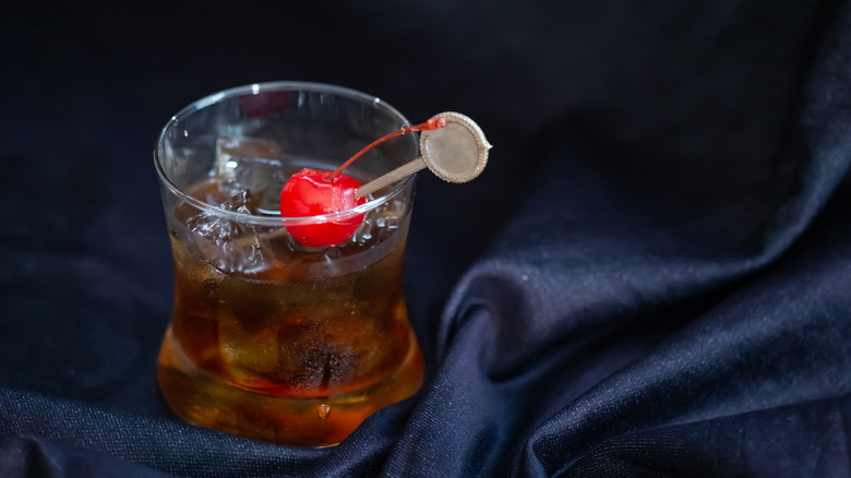 Dark brown cola cocktail with maraschino cherry in short glass sitting on dark blue velvet fabric