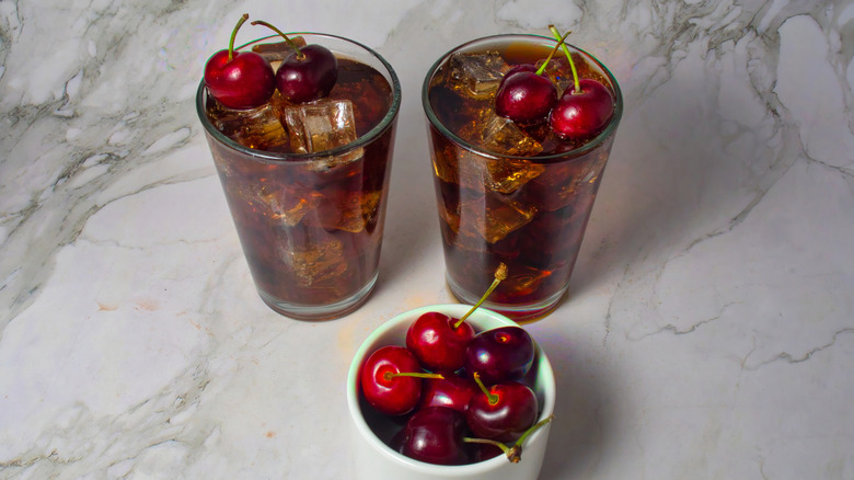 Two glasses of Roy Rogers mocktail garnished with black cherries next to white ramekins of cherries