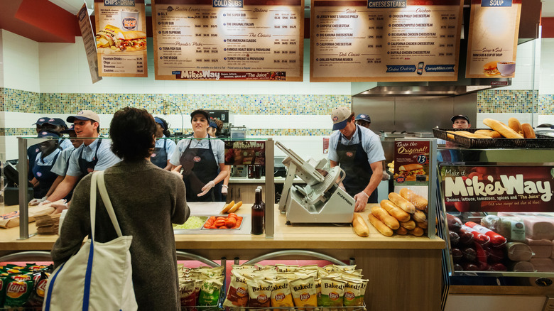 A customer stands in front of Jersey Mike's employees as they slice her proteins