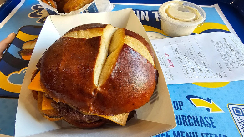 A Culver's Fried Pickle Pub Burger on a pretzel bun with some pickle chips and sauce on a tray