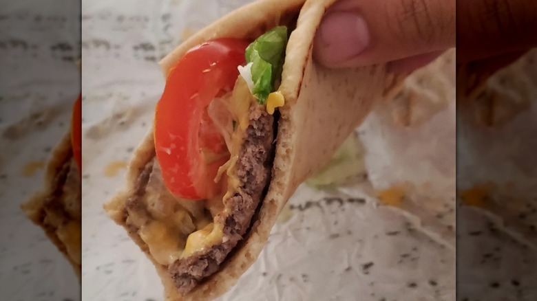 A person holding a Del Taco Burgertaco with a bite out of it showing a burger patty, lettuce, and tomato in a soft pita