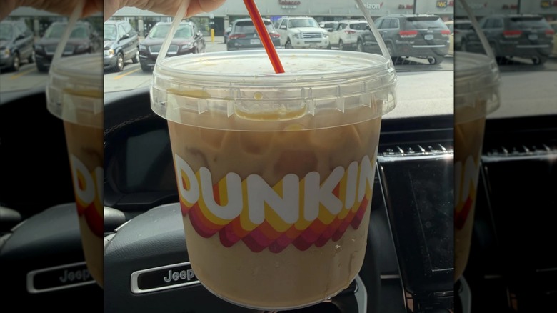 A Dunkin' Donuts branded bucket full of iced coffee being held up in front of a Jeep steering wheel