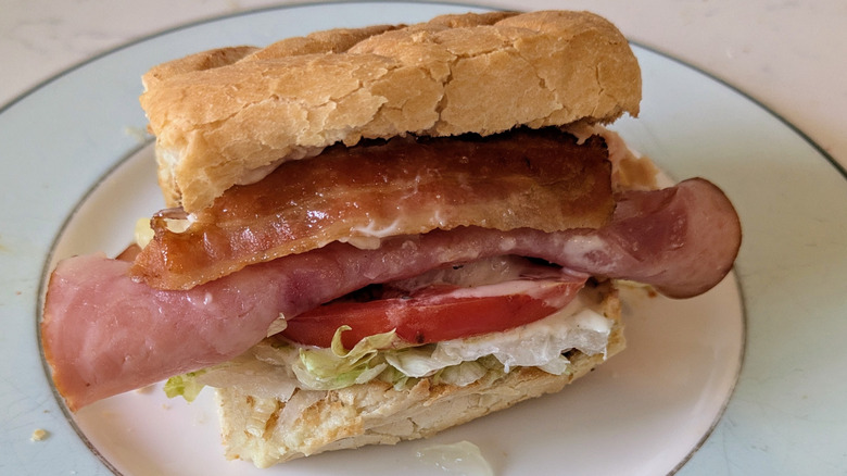 A toasted white sub hoagie roll is topped with shredded green lettuce, tomato slices, ham, and bacon on a white and blue plate.