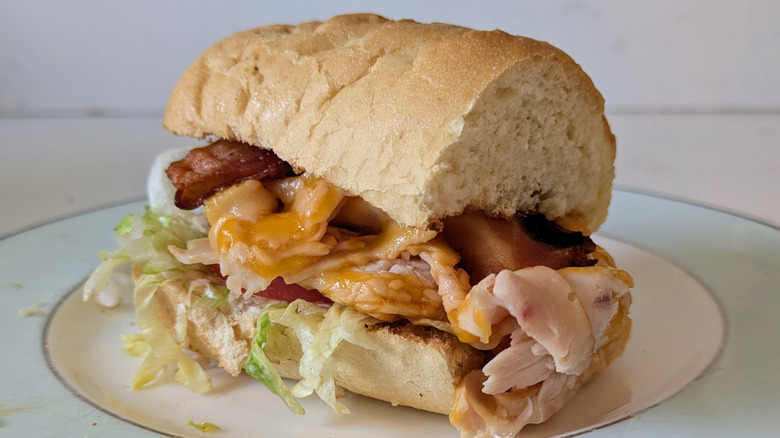 A toasted white sub hoagie roll is served on a blue and white plate, topped with shredded lettuce, tomato, cheddar cheese, bacon, and turkey