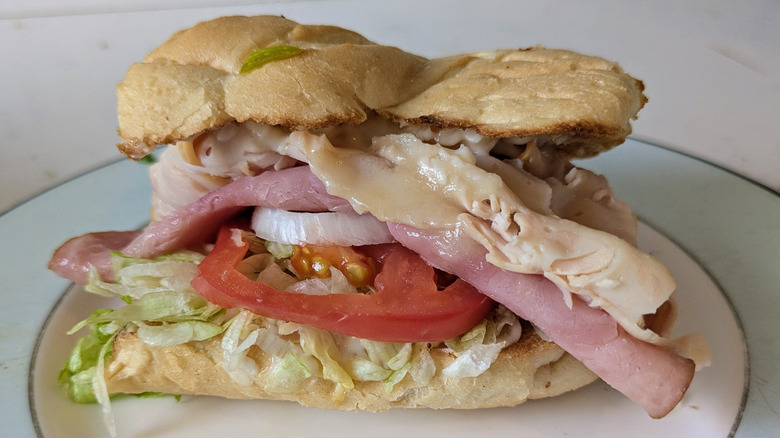 A white toasted sub hoagie roll is sliced in half and topped with shredded green lettuce, a sliced red tomato, sliced white onions, ham, turkey, and cheese on a blue and white plate